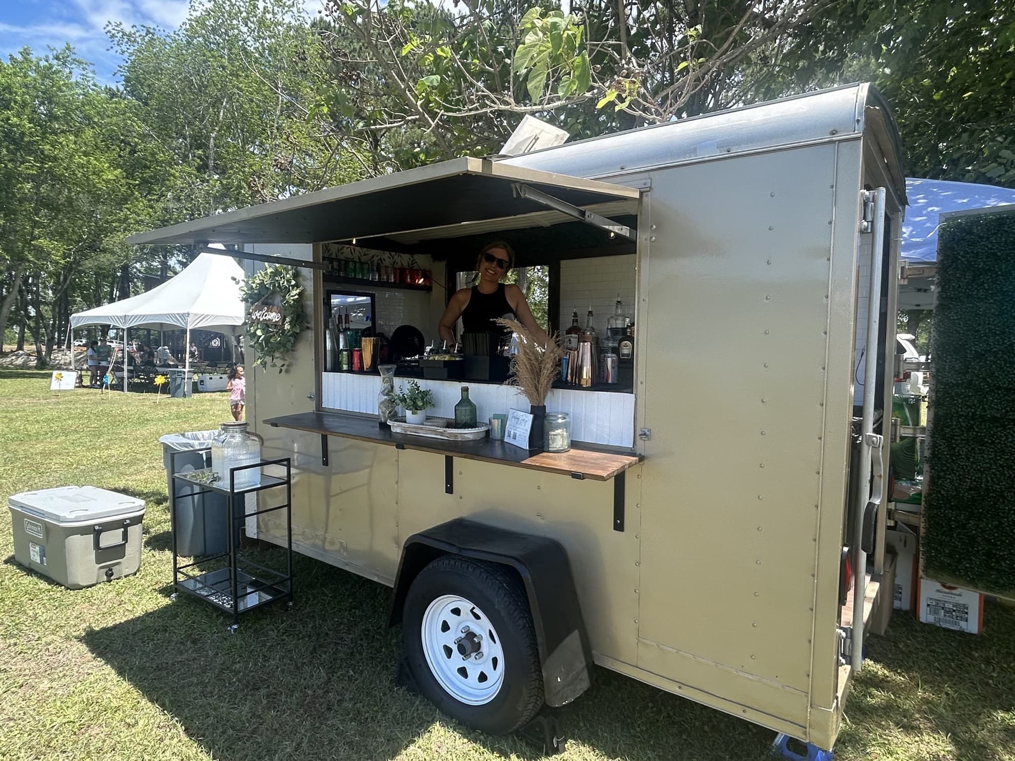 Bartender working the window of The Blair trailer at an outdoor festival, with tents and a cooler on the grass.