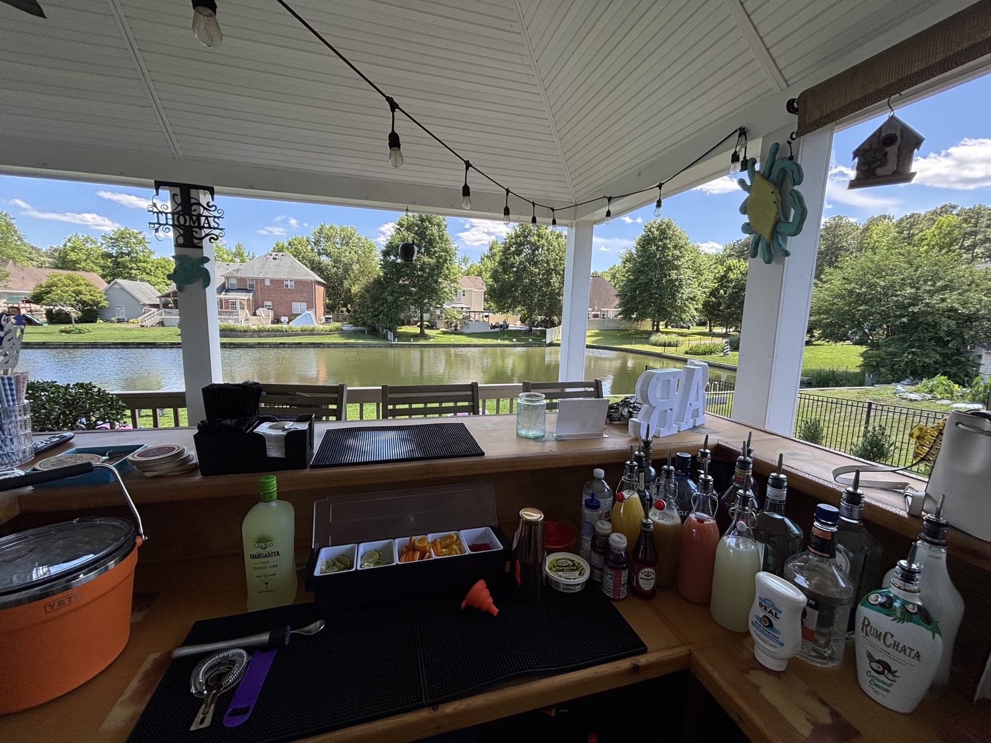 A bar station with bottles and mixers set up in a backyard gazebo overlooking a pond.