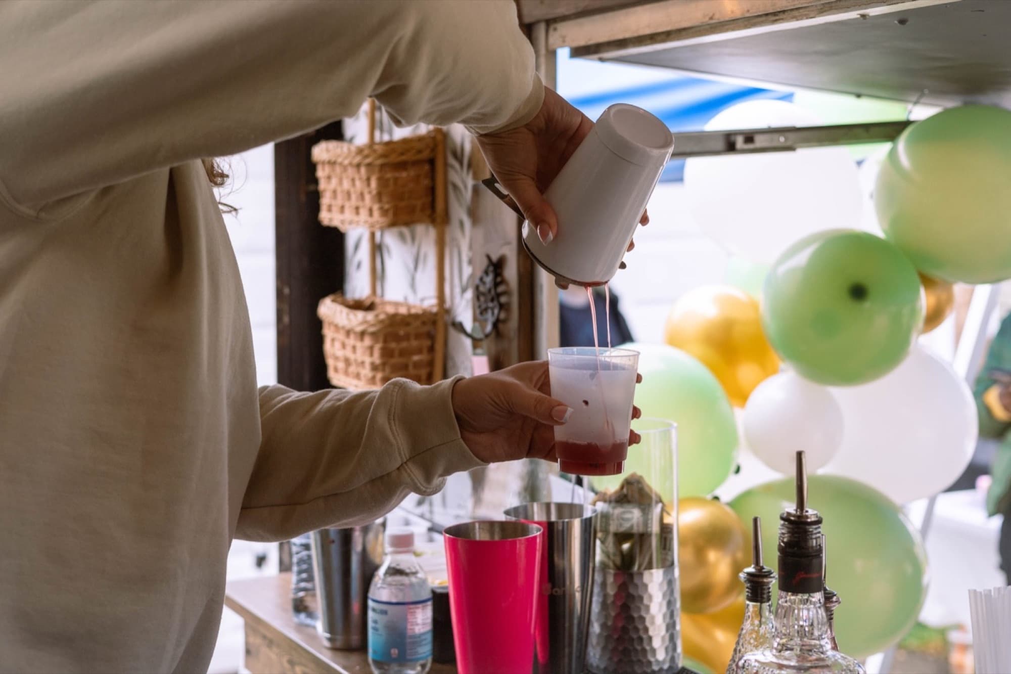 A bartender pours a red cocktail from the shaker inside The Blair, with green and gold balloons outside.