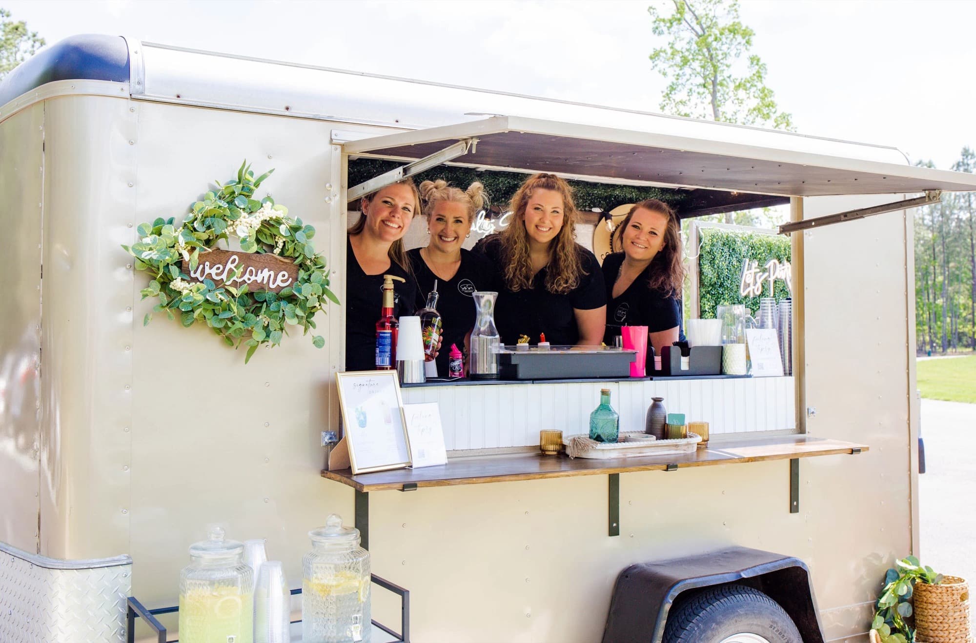 Four Salty Spirits bartenders smile from the open window of The Blair, with its welcome wreath and lemonade dispensers.